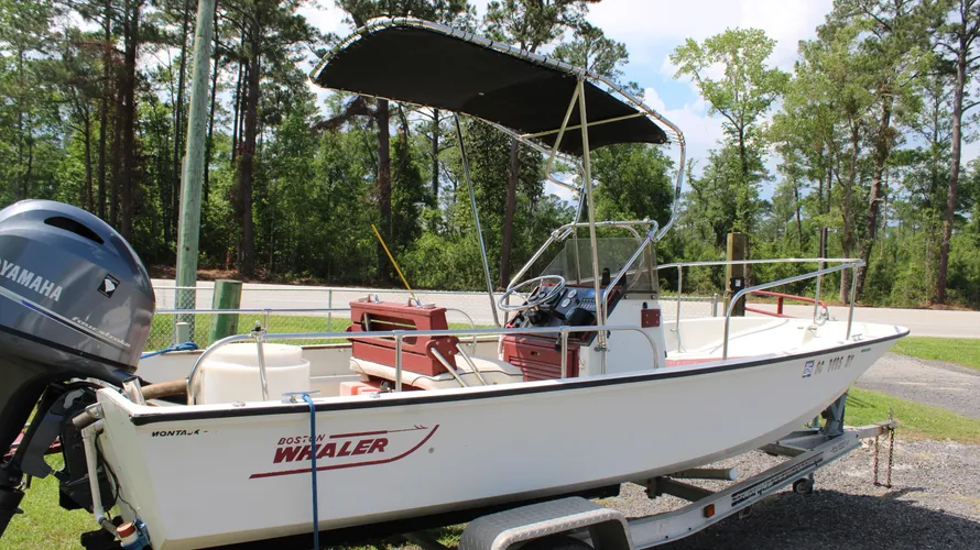 1982 Boston Whaler 17 Montauk center console boat with Yamaha 90hp outboard engine, featuring a bimini top and seating, parked on a trailer in a wooded area.