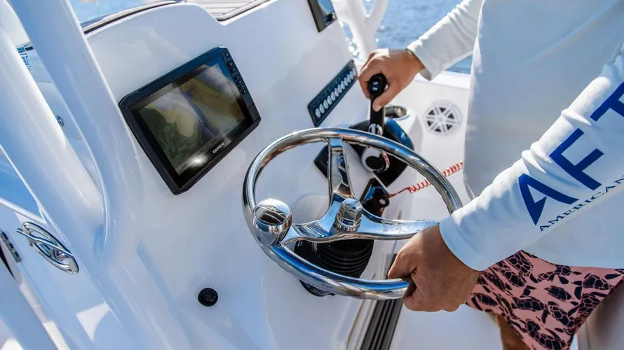 Boat steering console with a stainless steel wheel, GPS display, and throttle control, featuring a person in a long-sleeve shirt navigating a 2025 Tidewater Boats 198CC on water.